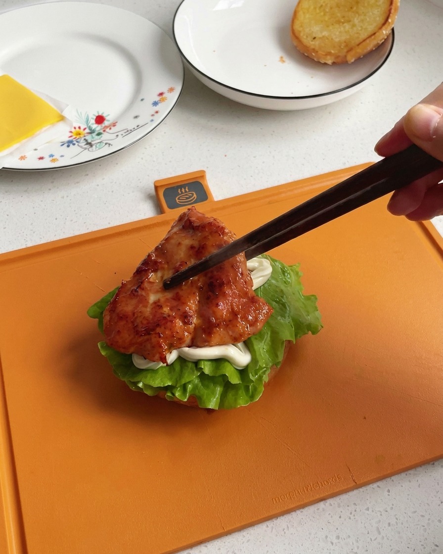 Une main utilisant des baguettes en bois sombre pour transférer un morceau de poulet poêlé, laqué et bien caramélisé, sur le montage du burger.