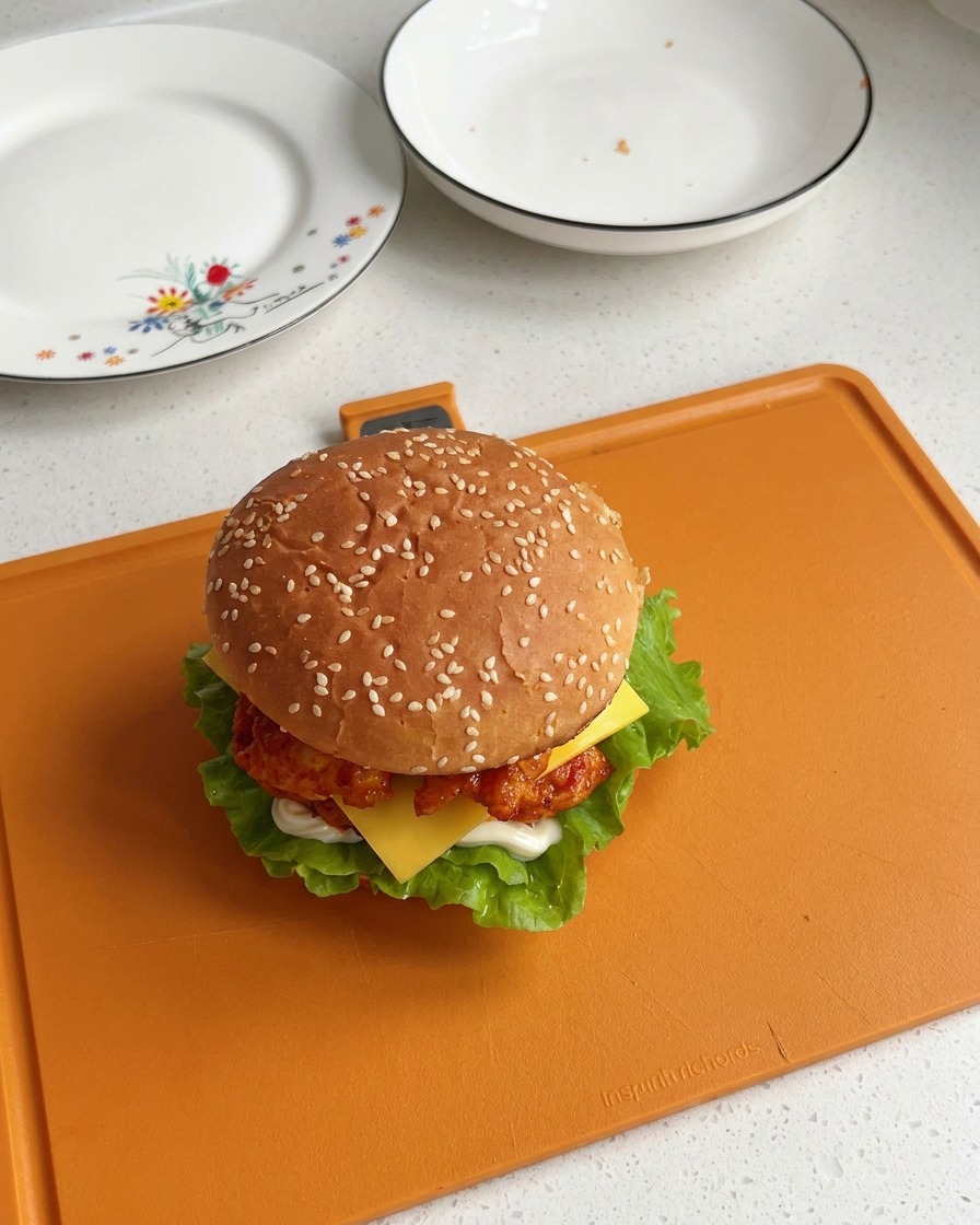Un burger au poulet dOrléans entièrement assemblé, coiffé de son pain au sésame, révélant de belles épaisseurs de salade, de poulet et une tranche de fromage fondante.