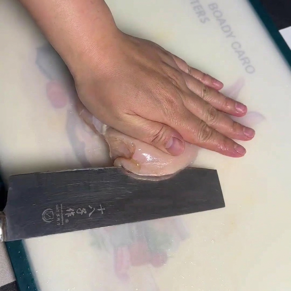 Une main maintenant un blanc de poulet cru à plat sur une planche à découper blanche tout en le coupant horizontalement avec un grand couteau de chef.