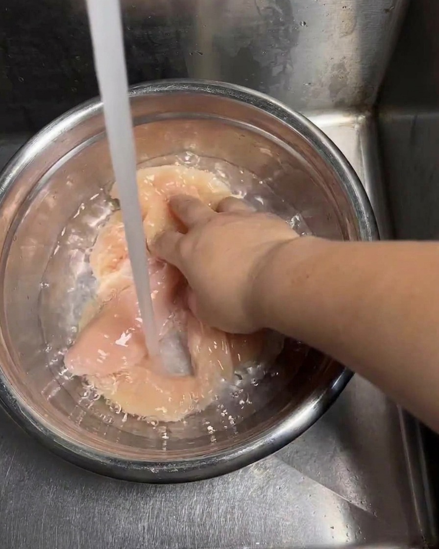 Un filet deau du robinet rinçant des escalopes de poulet crues dans un bol en inox pendant quune main remue les morceaux.