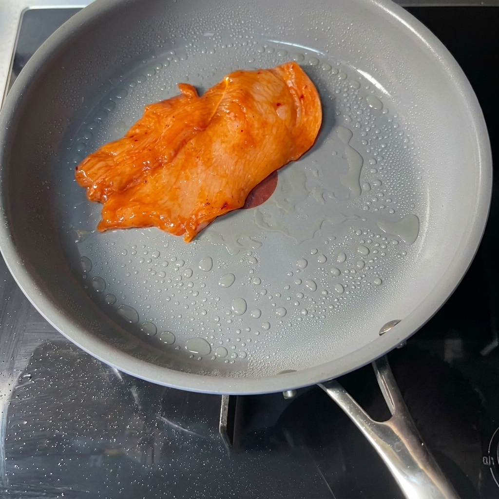 Une escalope de poulet marinée de couleur orange en train de cuire dans une poêle antiadhésive grise avec de petites bulles dhuile.