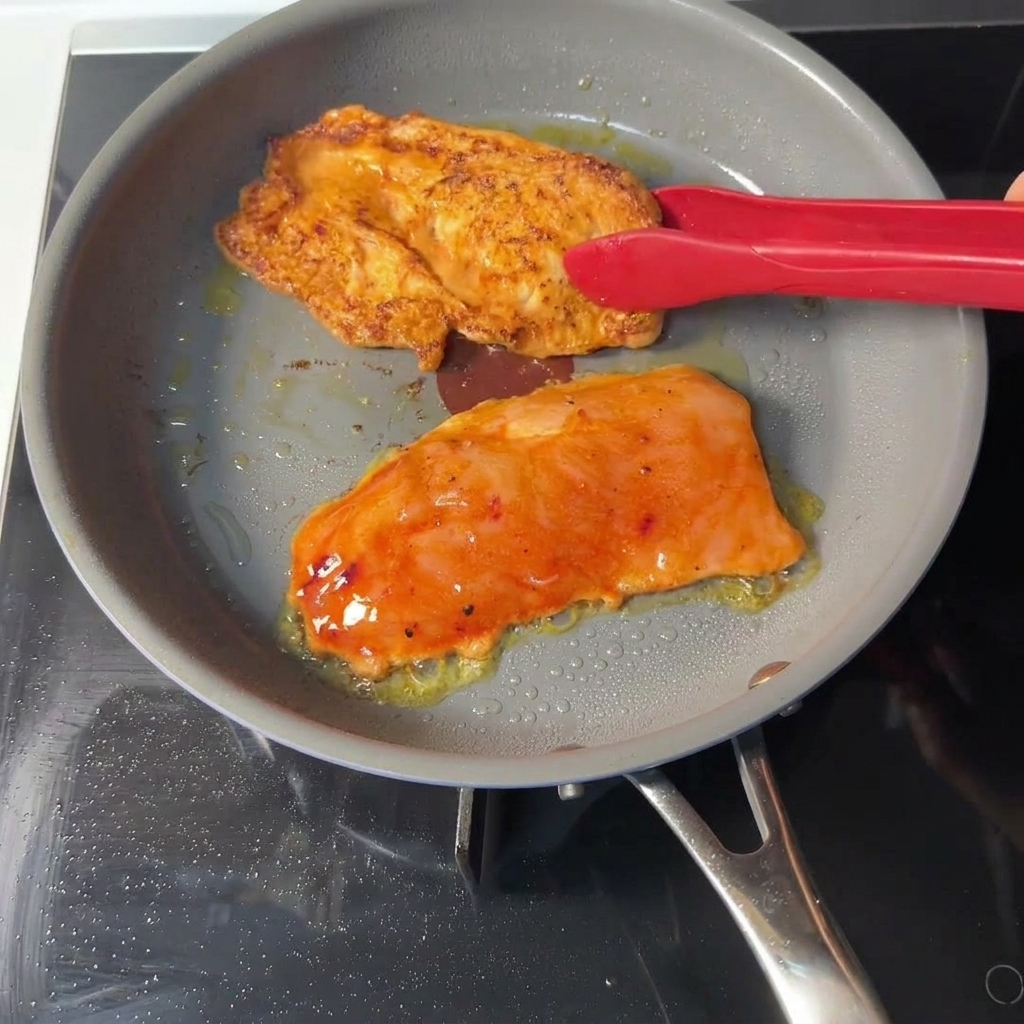 Une pince rouge retournant une escalope de poulet orange dans une poêle, révélant une face saisie et bien dorée.
