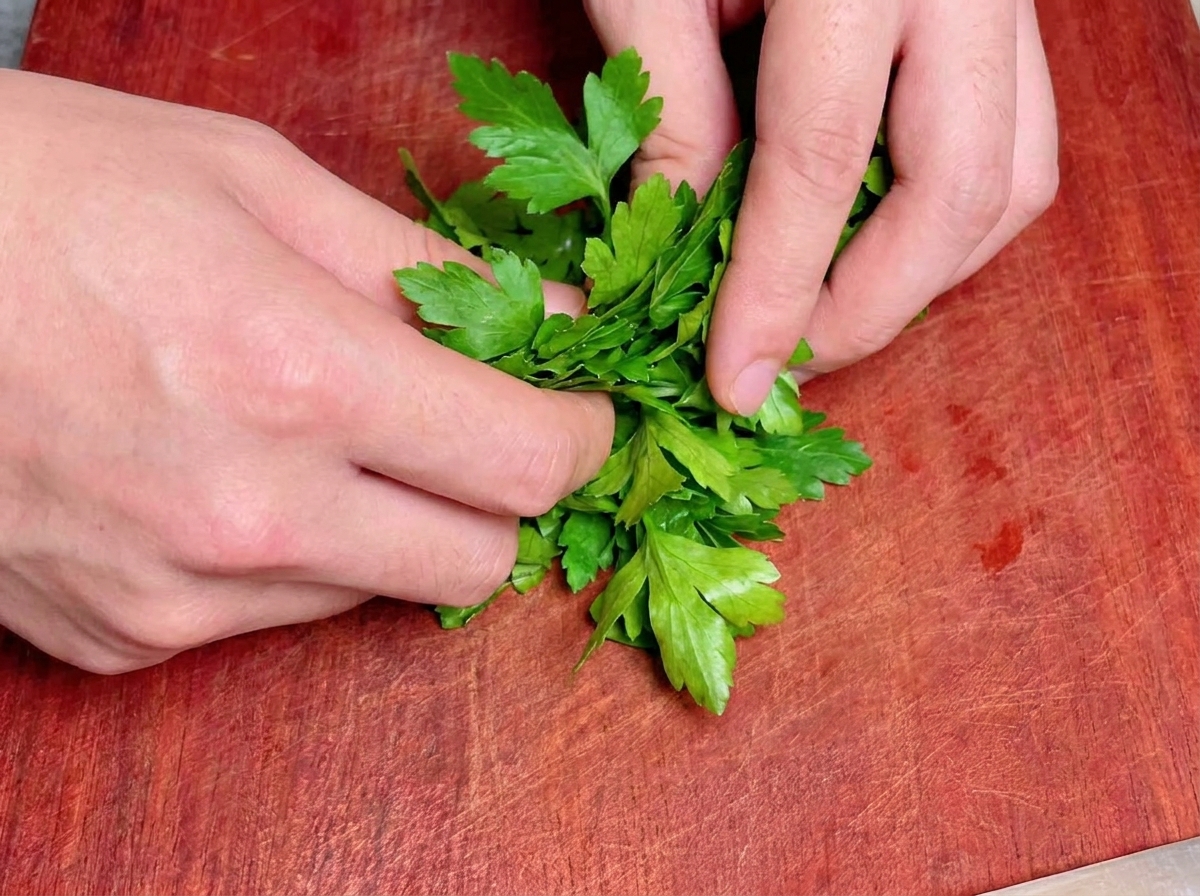 Deux mains enroulant serré un bouquet de persil plat vert frais sur une planche à découper en bois brun-rouge.