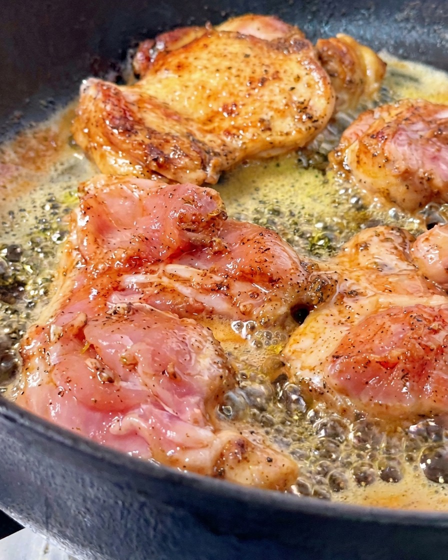 Des hauts de cuisse de poulet cuisant dans du beurre frémissant dans une poêle en fonte, un morceau étant retourné pour révéler une croûte dorée et croustillante.
