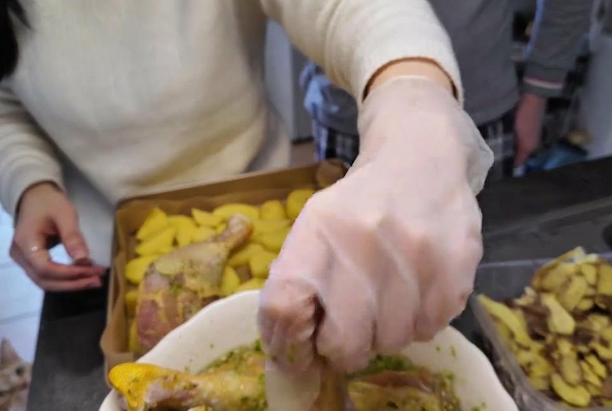 Une main gantée soulevant un pilon de poulet cru enrobé de marinade aux herbes vertes dun bol blanc.