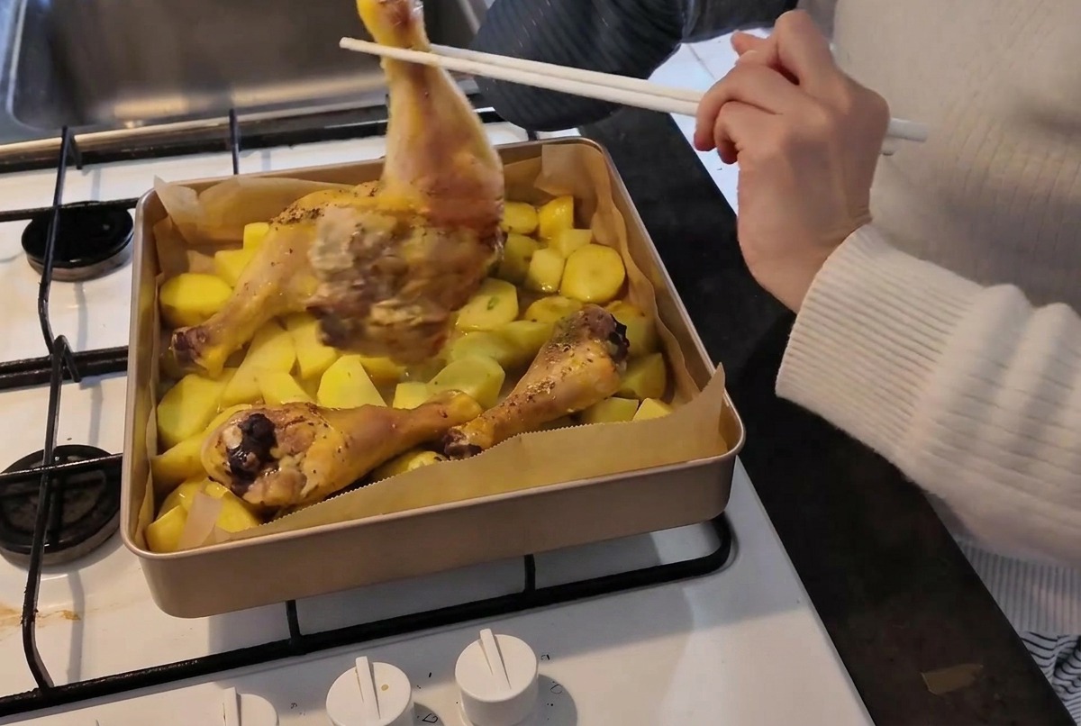 Une personne avec un gant de cuisine utilise des baguettes pour retourner une cuisse de poulet mi-cuite dans un plat chaud rempli de pommes de terre.