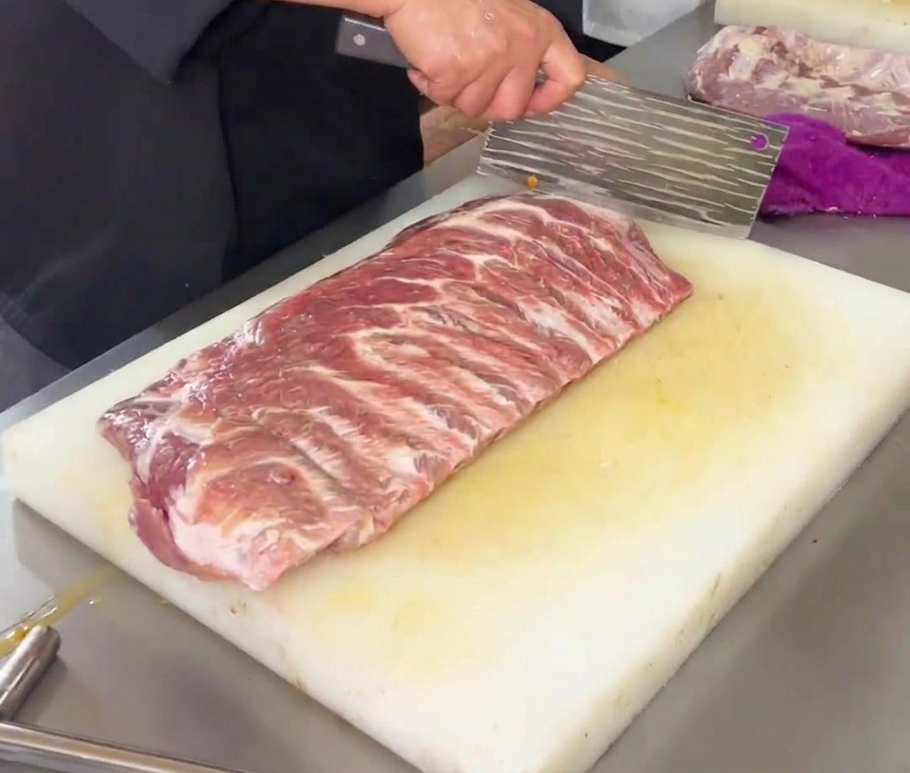 Un chef utilisant un grand couperet de cuisine pour entailler un motif en losange sur des travers de porc crus posés sur une planche à découper blanche.