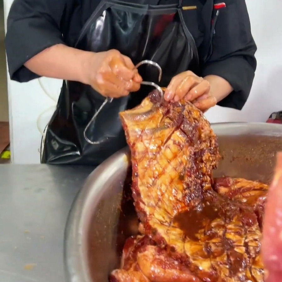 Un chef insérant un grand crochet métallique dans le coin supérieur dun travers de porc enrobé de marinade sombre au-dessus dun bol en inox.
