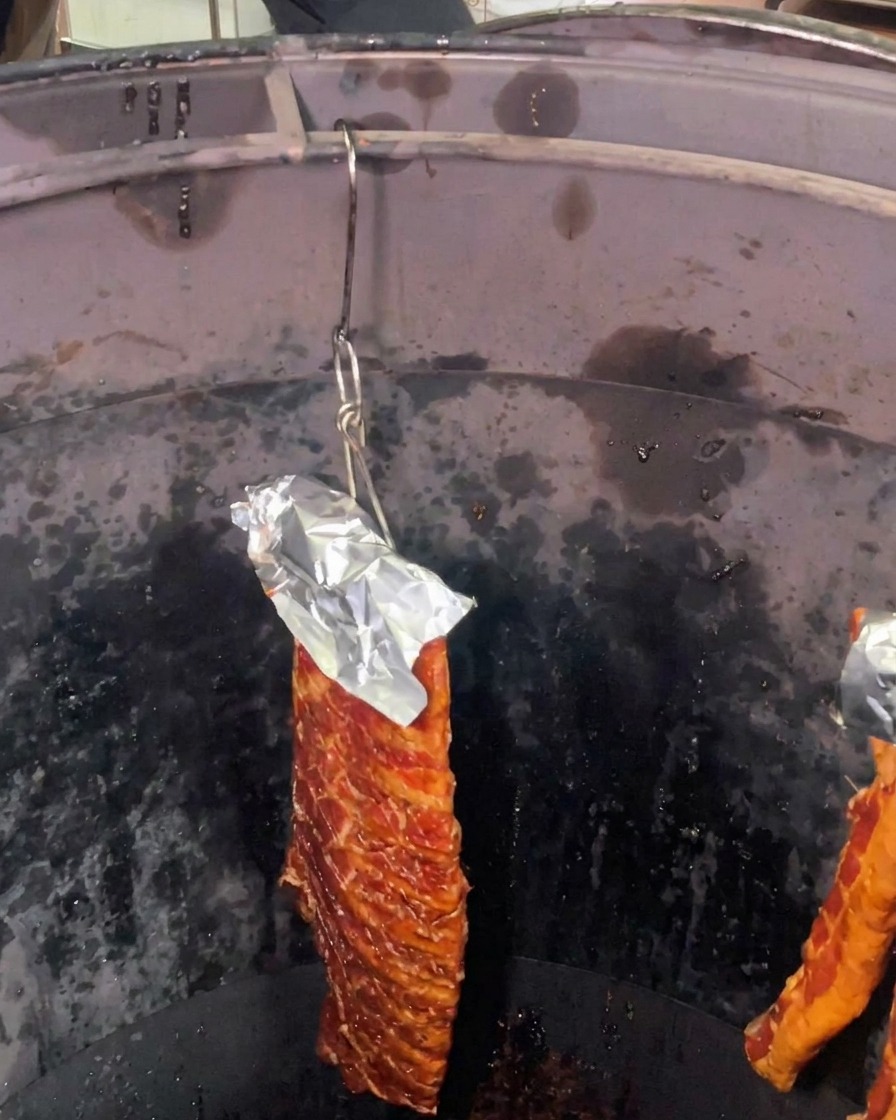 Un morceau de travers de porc mariné, protégé par de laluminium sur le dessus, suspendu à lintérieur dun grand four de rôtissage cylindrique sombre.