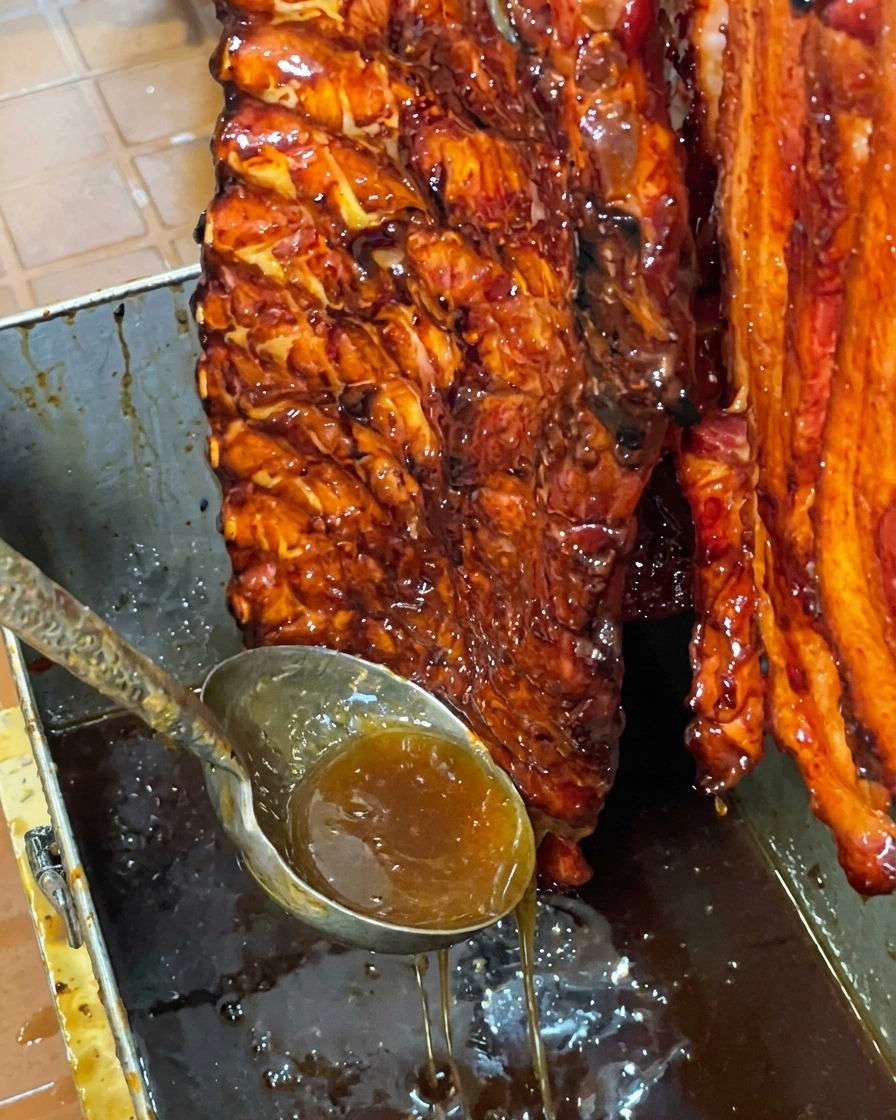 Un travers de porc laqué et brillant suspendu verticalement au-dessus dun bac en métal alors que lexcès de sirop ségoutte.