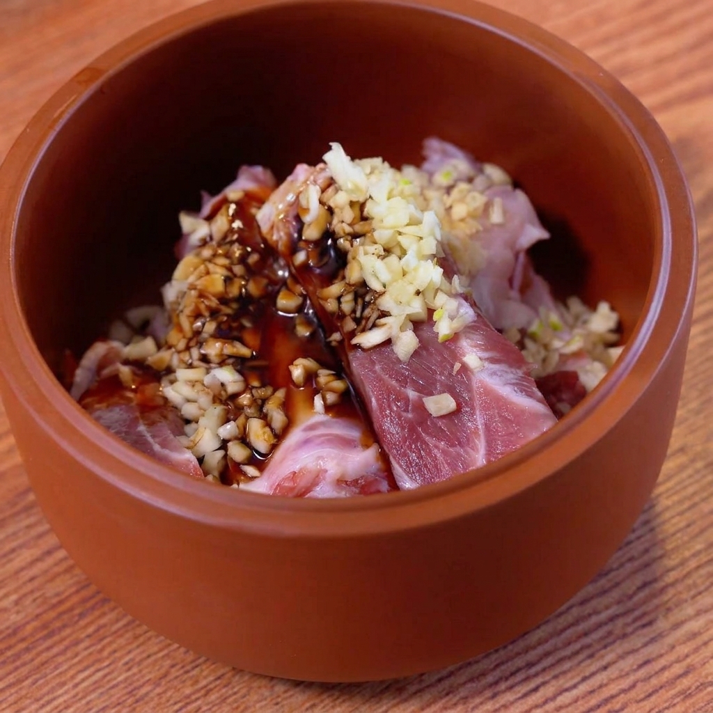 Travers de porc crus dans un bol en terre cuite marron, recouverts dail haché et de sauces sombres.