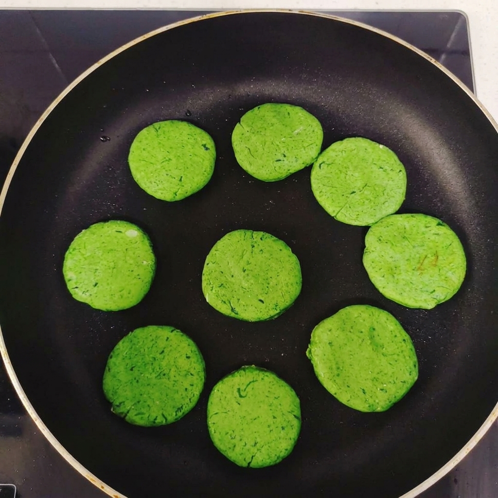 Plusieurs disques de galettes vertes disposés en cercle dans une poêle antiadhésive noire avant la cuisson.