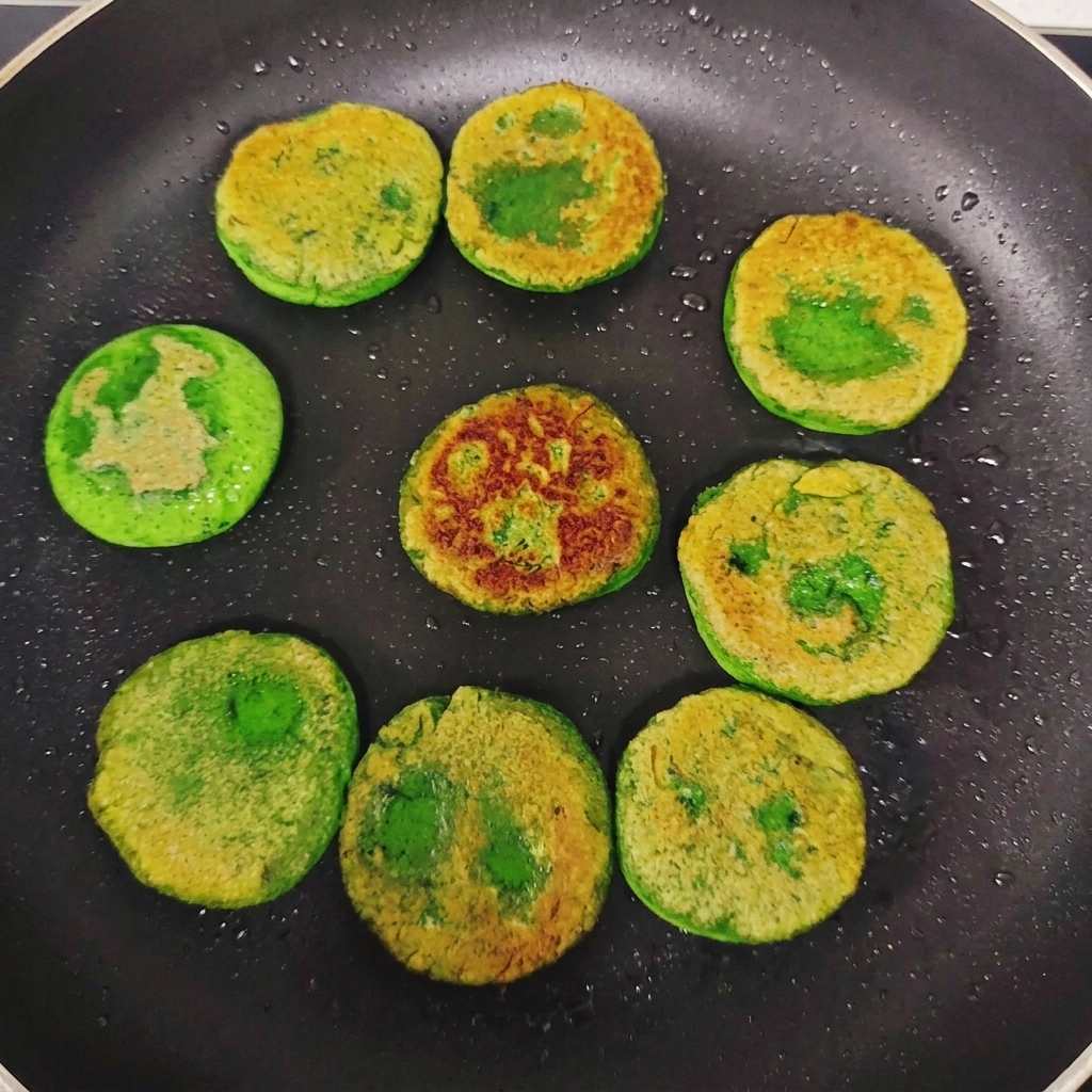 Galettes de légumes vertes retournées dans une poêle, montrant des zones dorées et saisies sur le dessus.