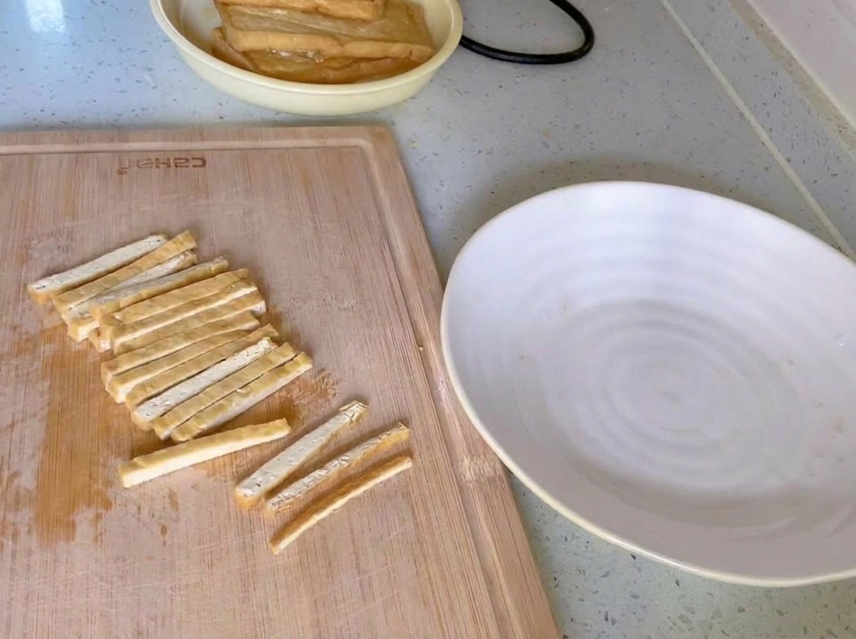 Tofu frit doré coupé en fines lanières uniformes sur une planche à découper en bois à côté dune assiette blanche.