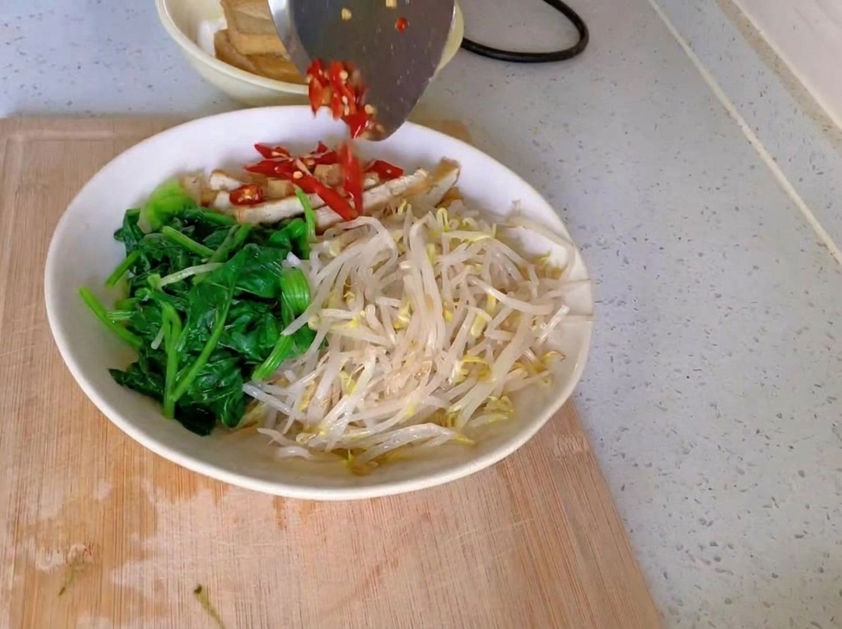 Un bol blanc contenant des sections bien distinctes dépinards verts, de germes de soja pâles et de lanières de tofu, surmontées dun tas de piments rouges hachés et dail émincé.