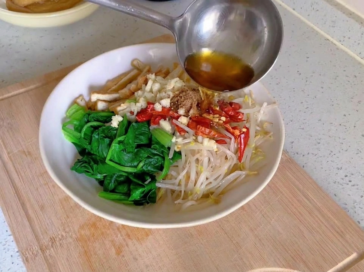 De lhuile fumante versée depuis une louche en métal sur de lail émincé et des piments rouges séchés disposés sur des légumes blanchis dans un bol blanc.