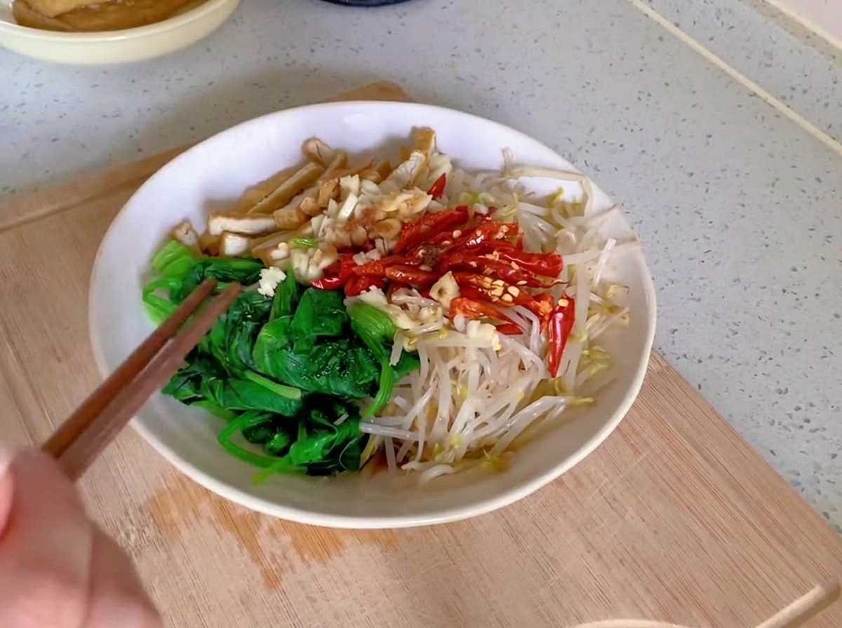 Une paire de baguettes en bois mélangeant des épinards verts, des germes de soja pâles, des lanières de tofu et des piments rouges dans un bol en céramique blanche.
