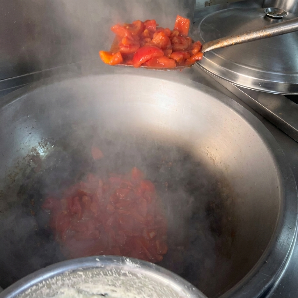 Une spatule en métal versant des tomates rouges concassées dans un grand wok en métal fumant contenant de lhuile chaude.