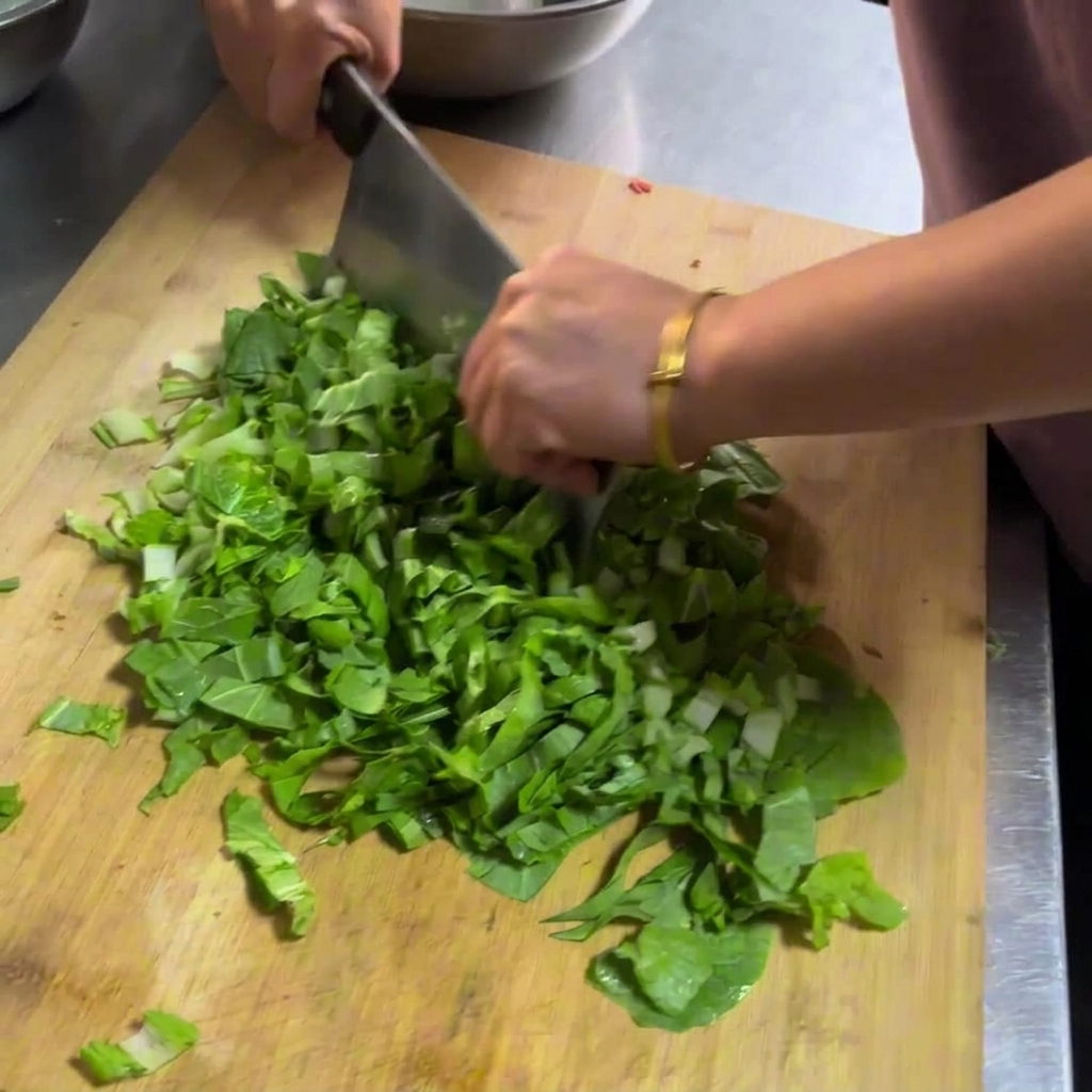 Mains utilisant un grand couperet en métal pour émincer des légumes verts sur une planche à découper en bois.