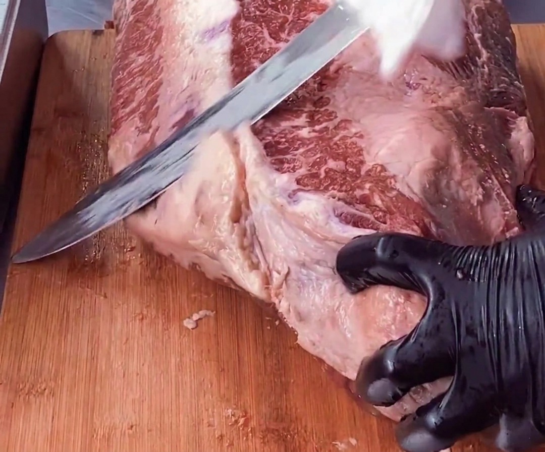 Une main gantée maintient un morceau de brisket tandis quun long couteau retire une épaisse couche de gras blanc de la viande rouge.
