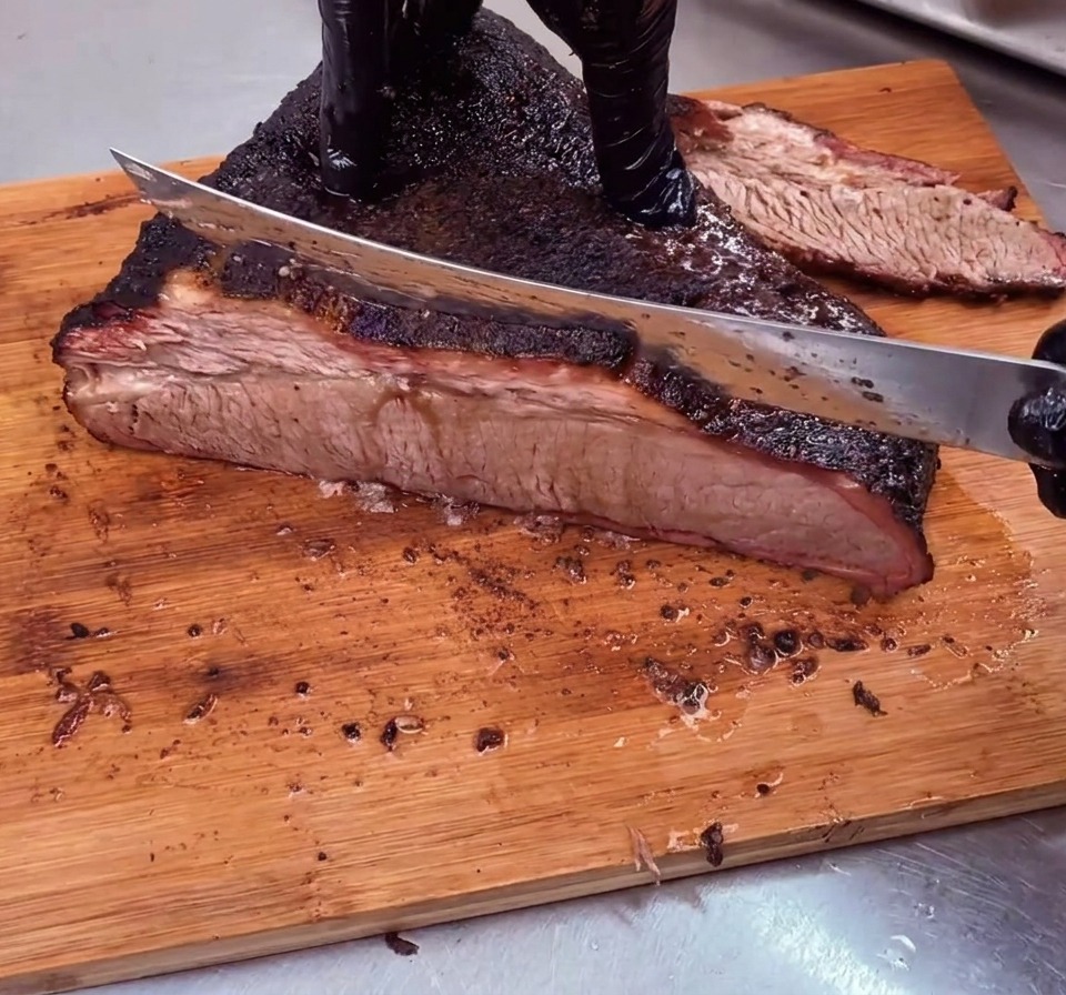 Un chef découpe en tranches fines un brisket de bœuf parfaitement fumé doté dune croûte sombre, perpendiculairement aux fibres, sur une planche en bois.
