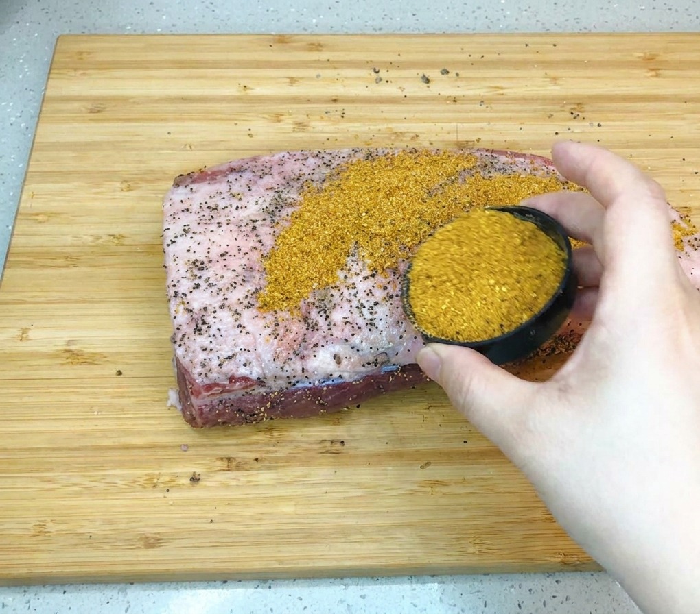 Une main versant un mélange dépices pour barbecue marron doré depuis un petit bol noir sur une grande poitrine de bœuf crue posée sur une planche en bois.