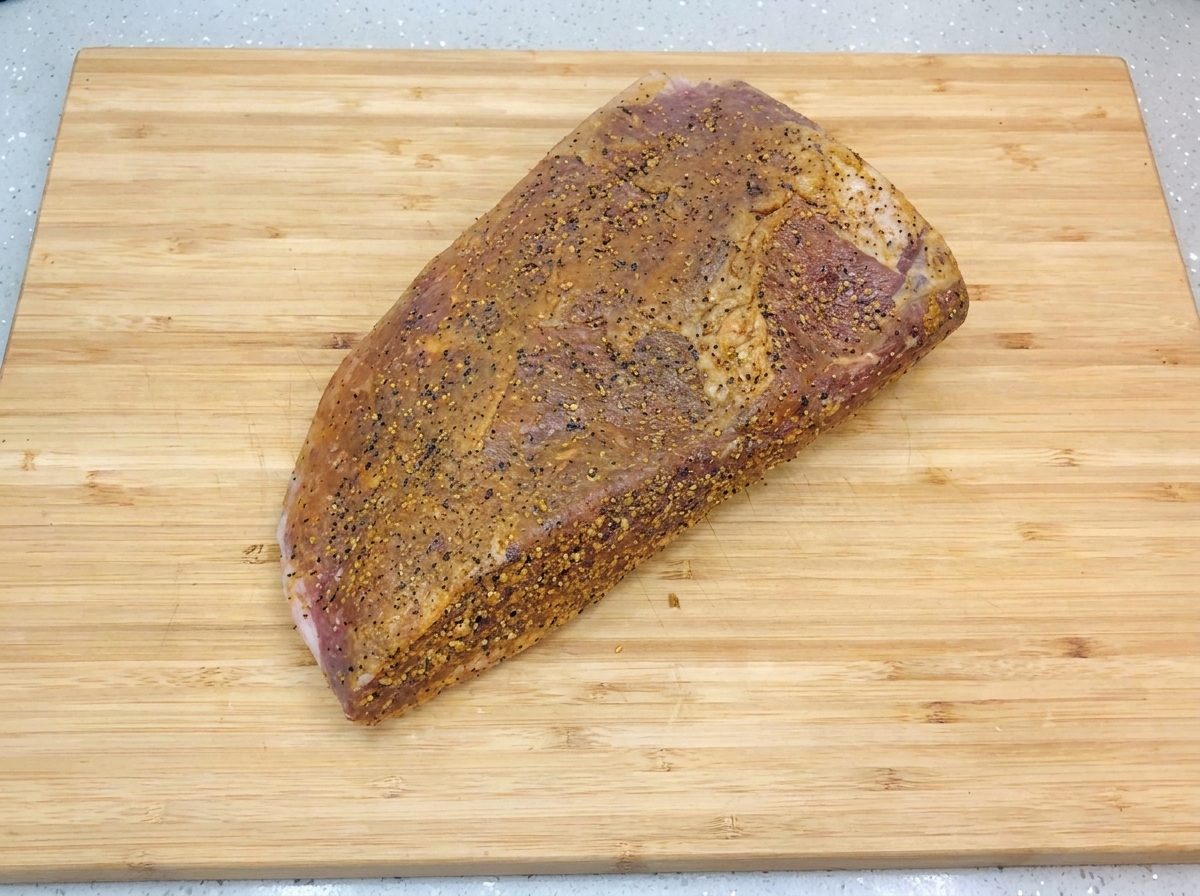 Une poitrine de bœuf marinée et assaisonnée reposant sur une planche à découper en bois clair, brillante sous une fine couche dhuile et parsemée de poivre noir moulu.