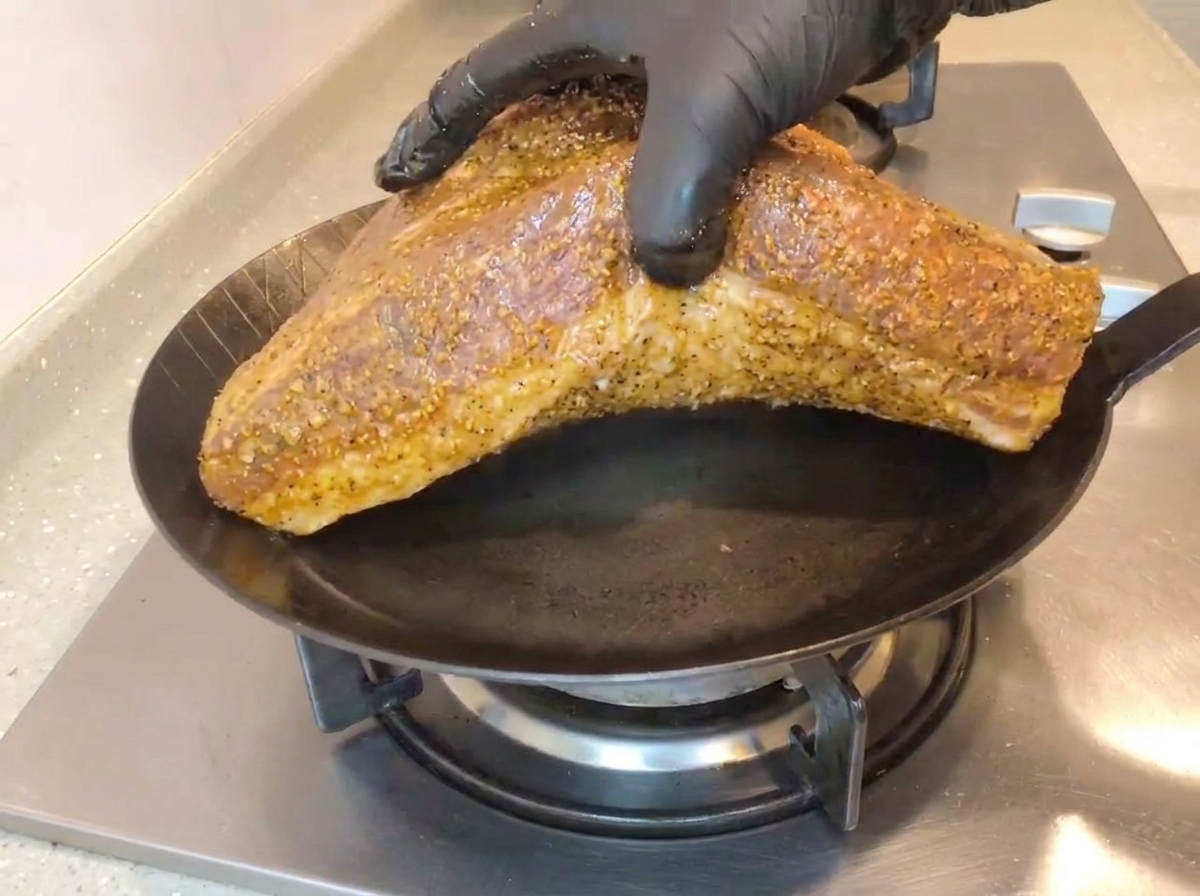 Une main avec un gant noir déposant une grande poitrine de bœuf assaisonnée, côté gras vers le bas, dans une poêle en fonte noire bien chaude sur une cuisinière à gaz.