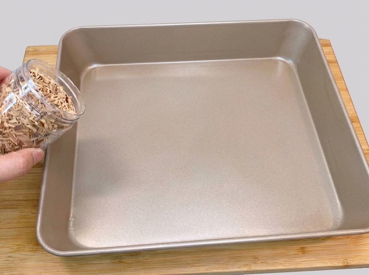 Une main versant des copeaux de bois de pêcher depuis un récipient en plastique directement au fond dun plat de cuisson carré de couleur dorée.