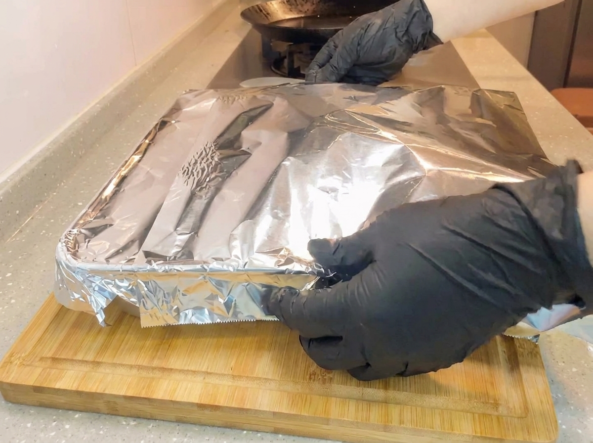 Des mains gantées rabattant fermement une feuille de papier daluminium sur un plat de cuisson carré posé sur une planche en bois.