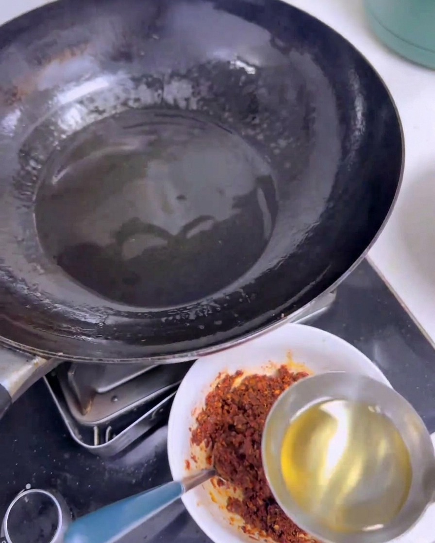 Une louche en inox versant de lhuile chaude et claire dans un bol blanc contenant un mélange de piments rouges à côté dun wok noir.