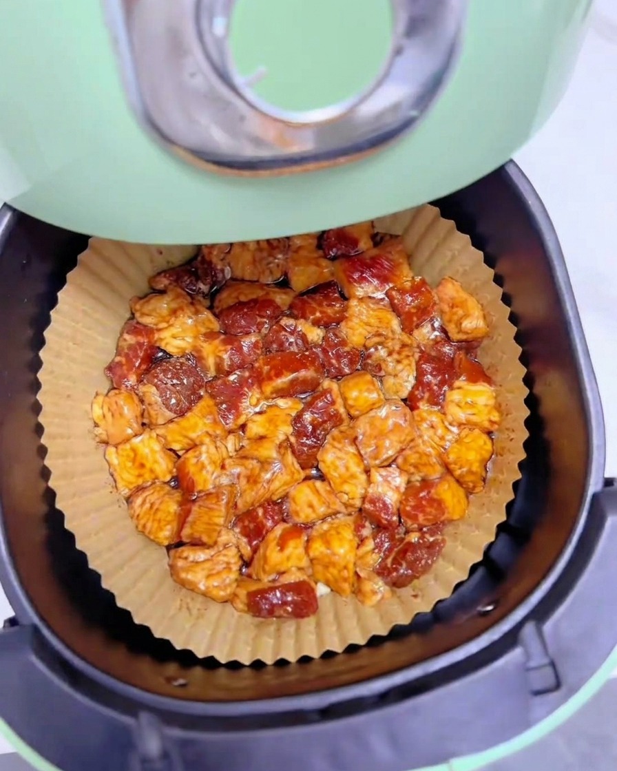 Morceaux de gras de bœuf marinés répartis uniformément en une seule couche sur du papier cuisson dans le panier dun air fryer vert.