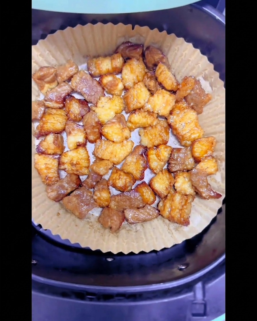 Morceaux de gras de bœuf partiellement dorés disposés sur une feuille de papier cuisson ronde dans le panier de lair fryer.