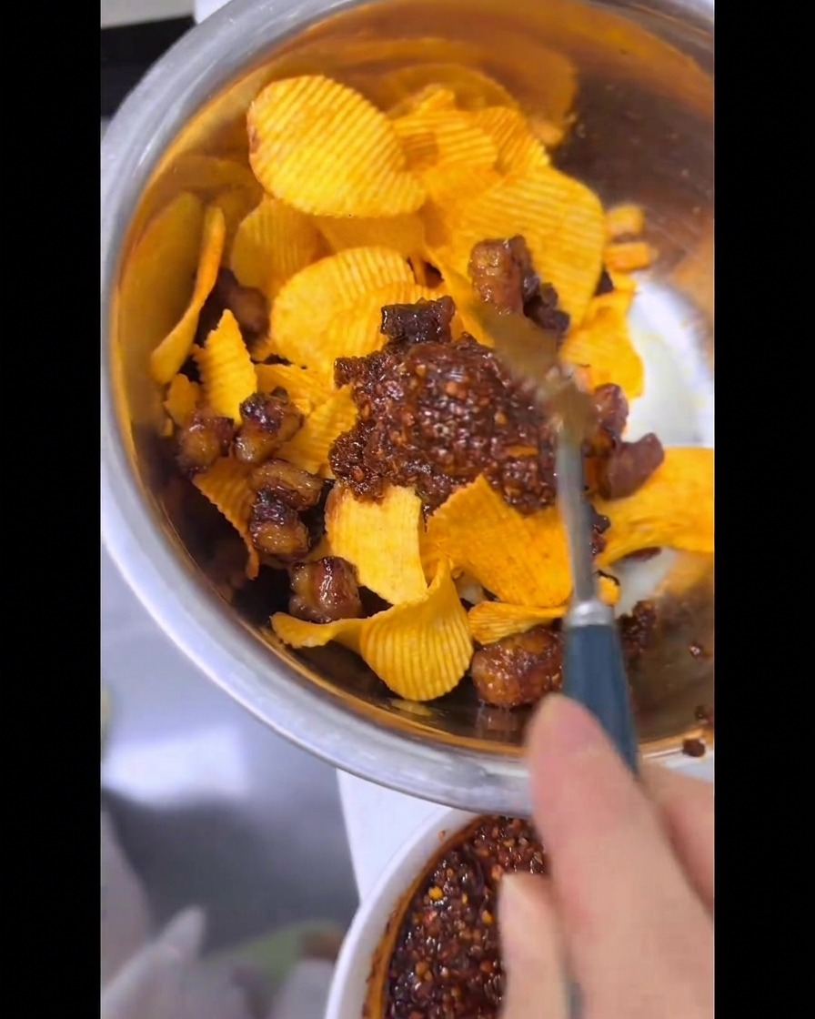 Une main utilisant une cuillère pour mélanger des chips ondulées, du gras de bœuf croustillant et une huile pimentée rouge vif dans un bol en métal.