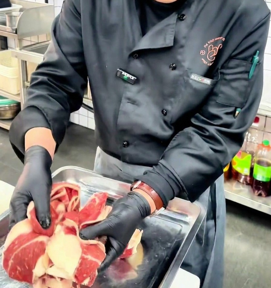 Un chef portant des gants noirs présente de fines tranches de poitrine de bœuf crue sur un plateau métallique.