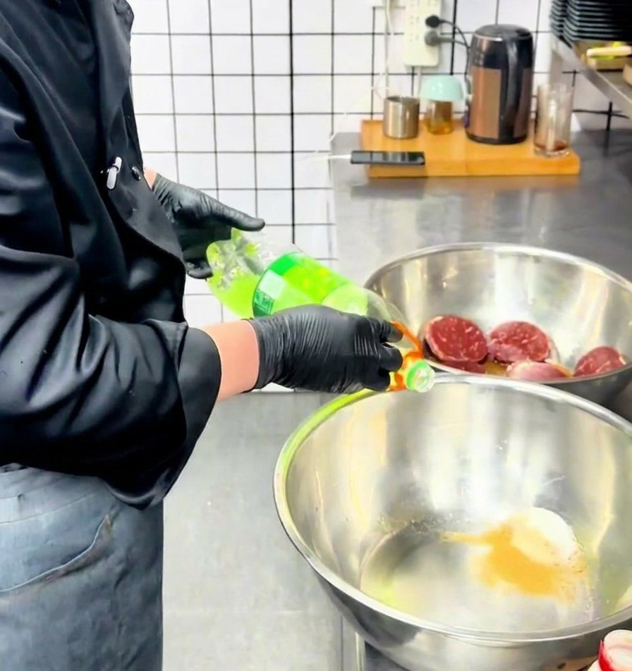 Un chef verse un liquide vert clair depuis une bouteille dans un bol en inox contenant les épices.