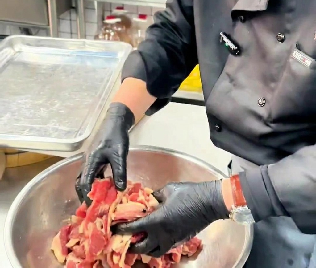 Des mains gantées remuent des tranches fines de bœuf enrobées dhuile dans un bol en métal.