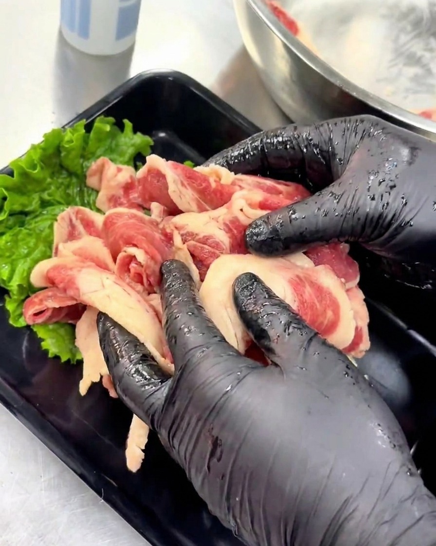 Des mains gantées disposent soigneusement les tranches de bœuf mariné sur un plateau noir décoré dune feuille de laitue.