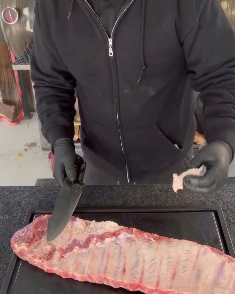 Une personne portant des gants noirs pare les morceaux de viande superflus et les bords irréguliers dun travers de porc cru à laide dun grand couteau de chef sur une planche à découper noire.