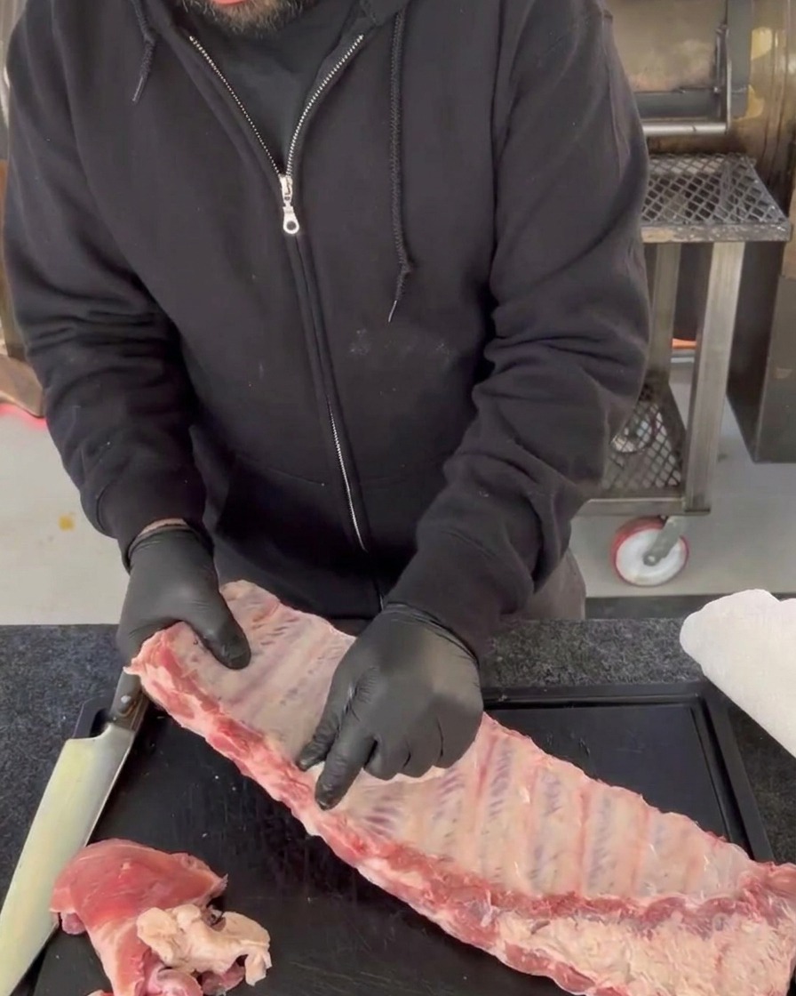 Des mains gantées décollent la membrane brillante et translucide accrochée aux os à larrière dun travers de porc cru sur une planche à découper.