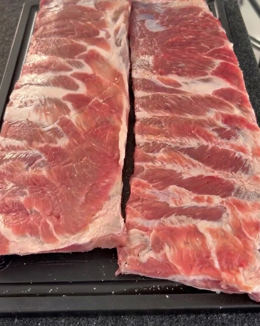 Deux travers de porc crus disposés côte à côte sur une planche à découper, présentant leur côté charnu recouvert de poivre concassé et de sel.