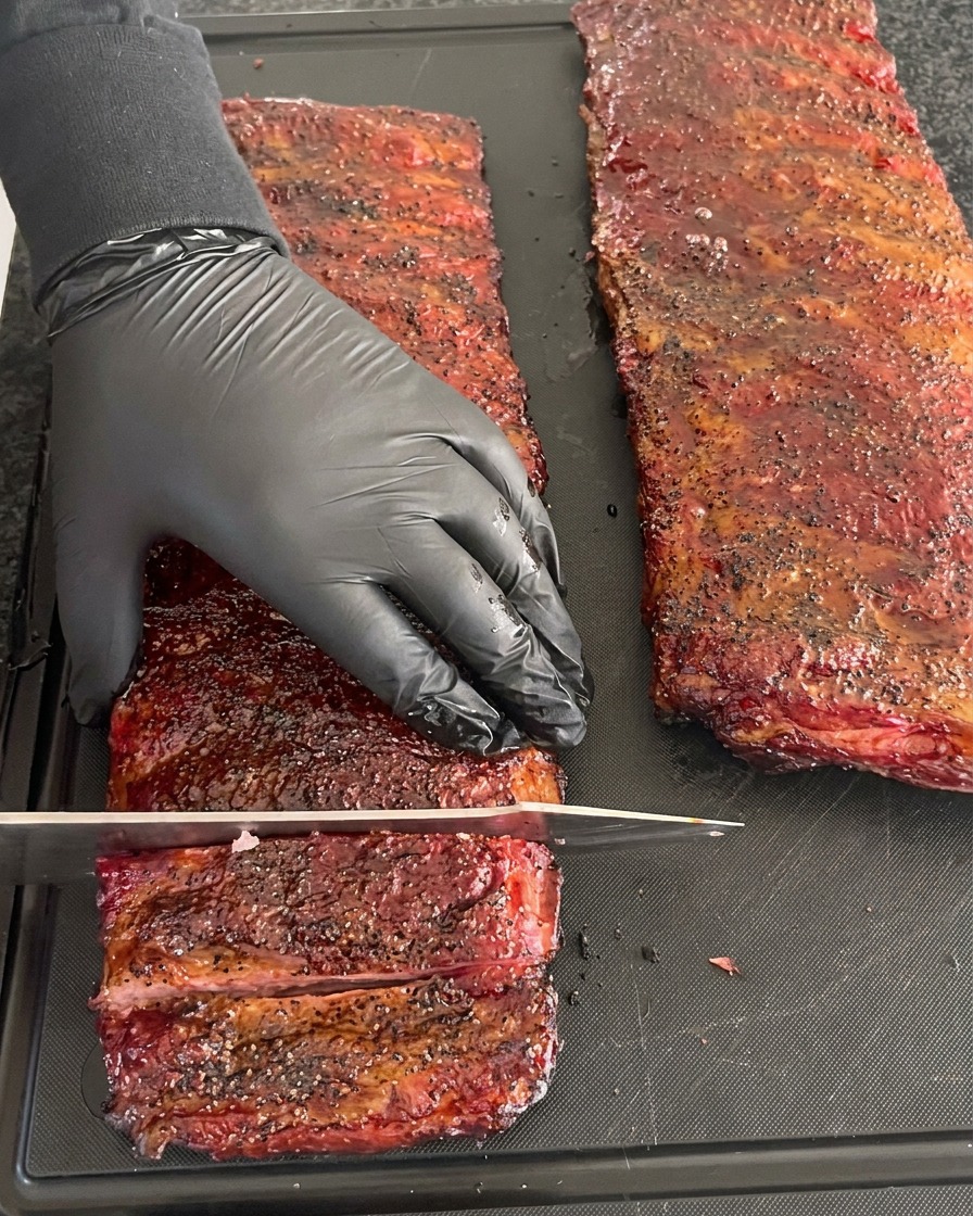 Une personne portant un gant noir maintient un travers de porc fumé à la croûte foncée tout en coupant les côtes individuellement avec un grand couteau.