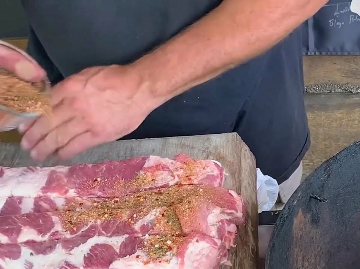 Une personne saupoudrant un mélange dépices sur de longues bandes de travers de porc crus sur une planche en bois.