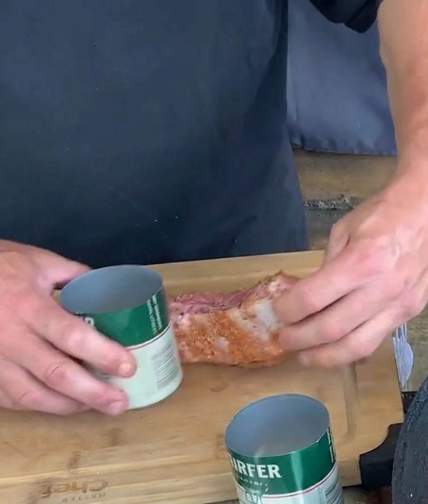 Des mains enroulant une bande de travers de porc assaisonnée autour dune canette en aluminium ouverte sur une planche à découper.
