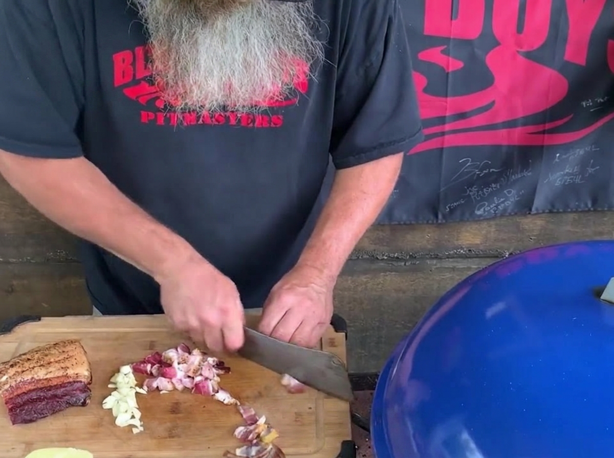 Un homme à barbe grise utilisant un grand couperet de cuisine pour hacher du bacon et des gousses dail sur une planche en bois à côté dun barbecue bleu.