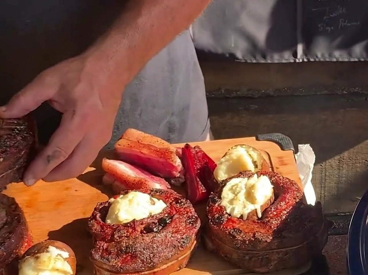 Une main plaçant une pomme de terre rôtie bien chaude et beurrée au centre dun panier de ribs cuit sur une planche à découper en bois.