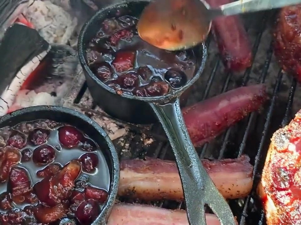 Une cuillère versant une riche sauce barbecue aux myrtilles depuis une petite casserole en fonte sur un panier de ribs farci sur le barbecue.