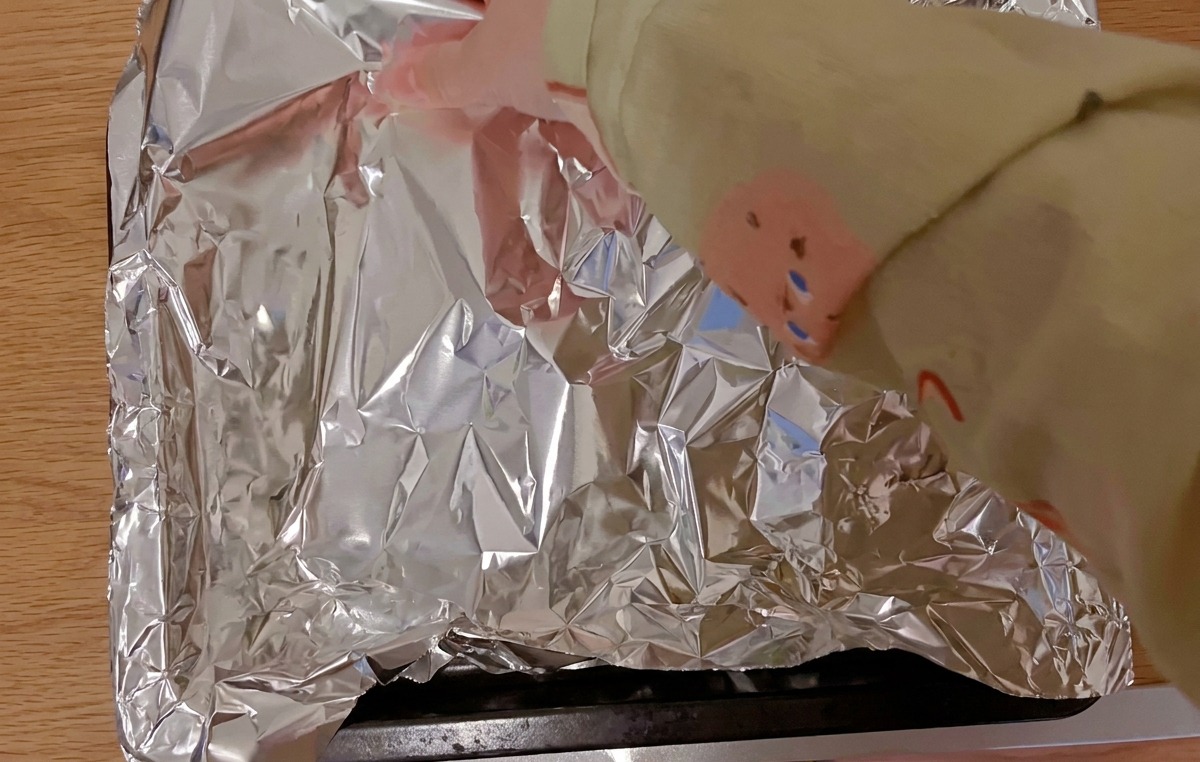 Des mains recouvrant les travers de porc marinés disposés sur une plaque de cuisson noire avec une feuille de papier daluminium brillant.