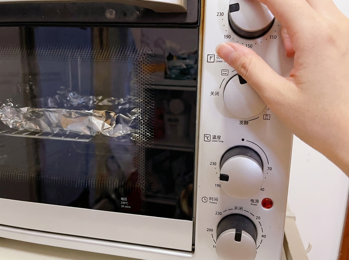 Gros plan dune main ajustant le bouton de température dun mini-four électrique blanc sur 230°C, avec les éléments emballés dans de laluminium visibles sur la grille.