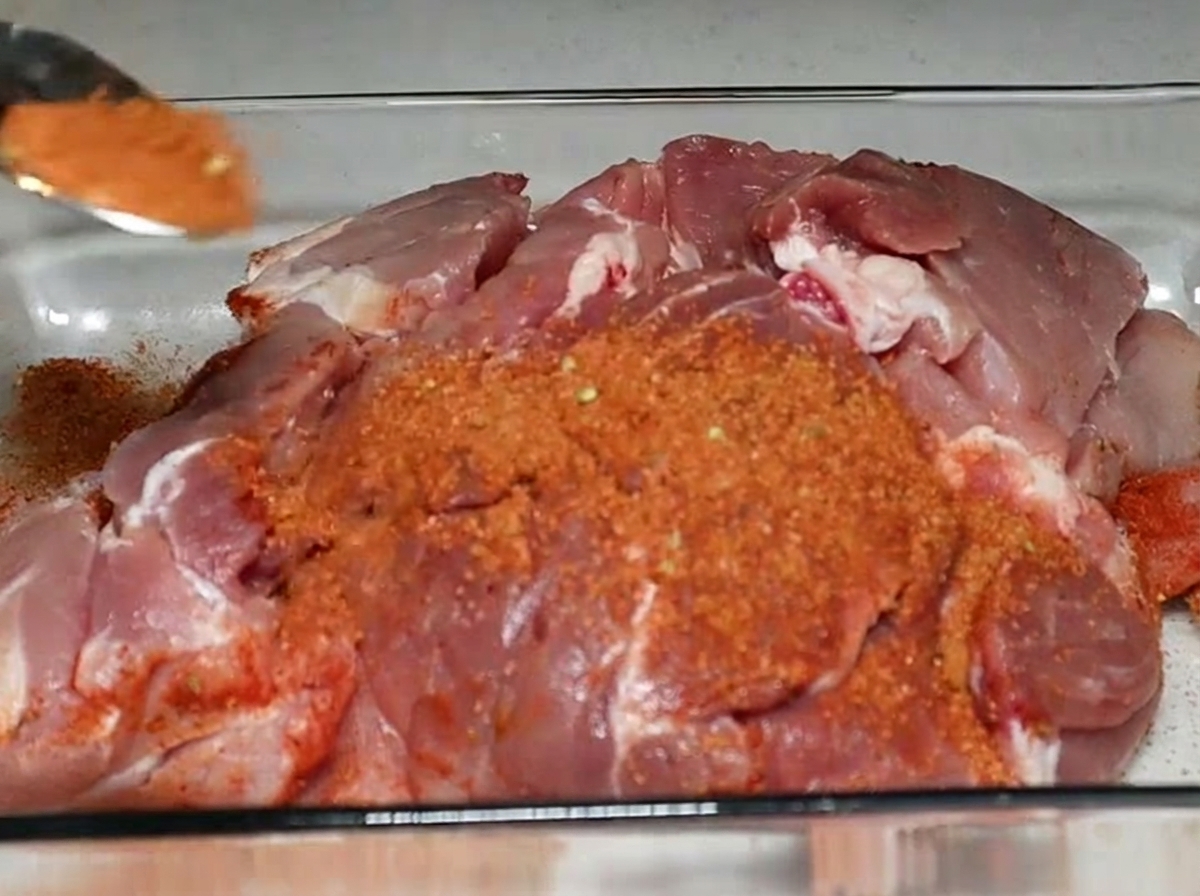 Une cuillérée de mélange dépices rougeâtres saupoudrée sur un grand morceau de viande de porc crue dans un plat en verre.